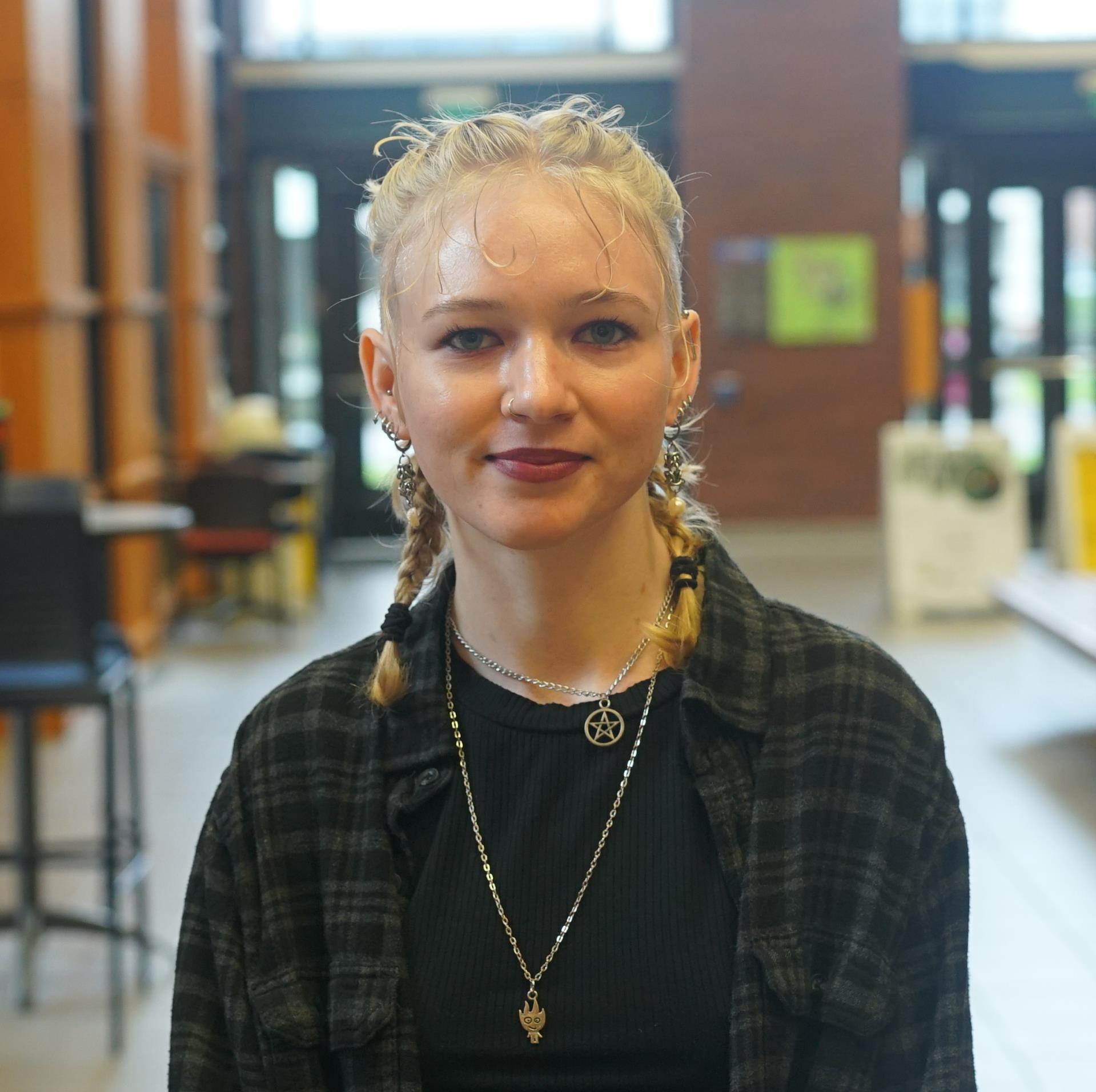 Portrait of Siobhan standing in the Heiner Foyer. They are smiling with blond hair in french braids