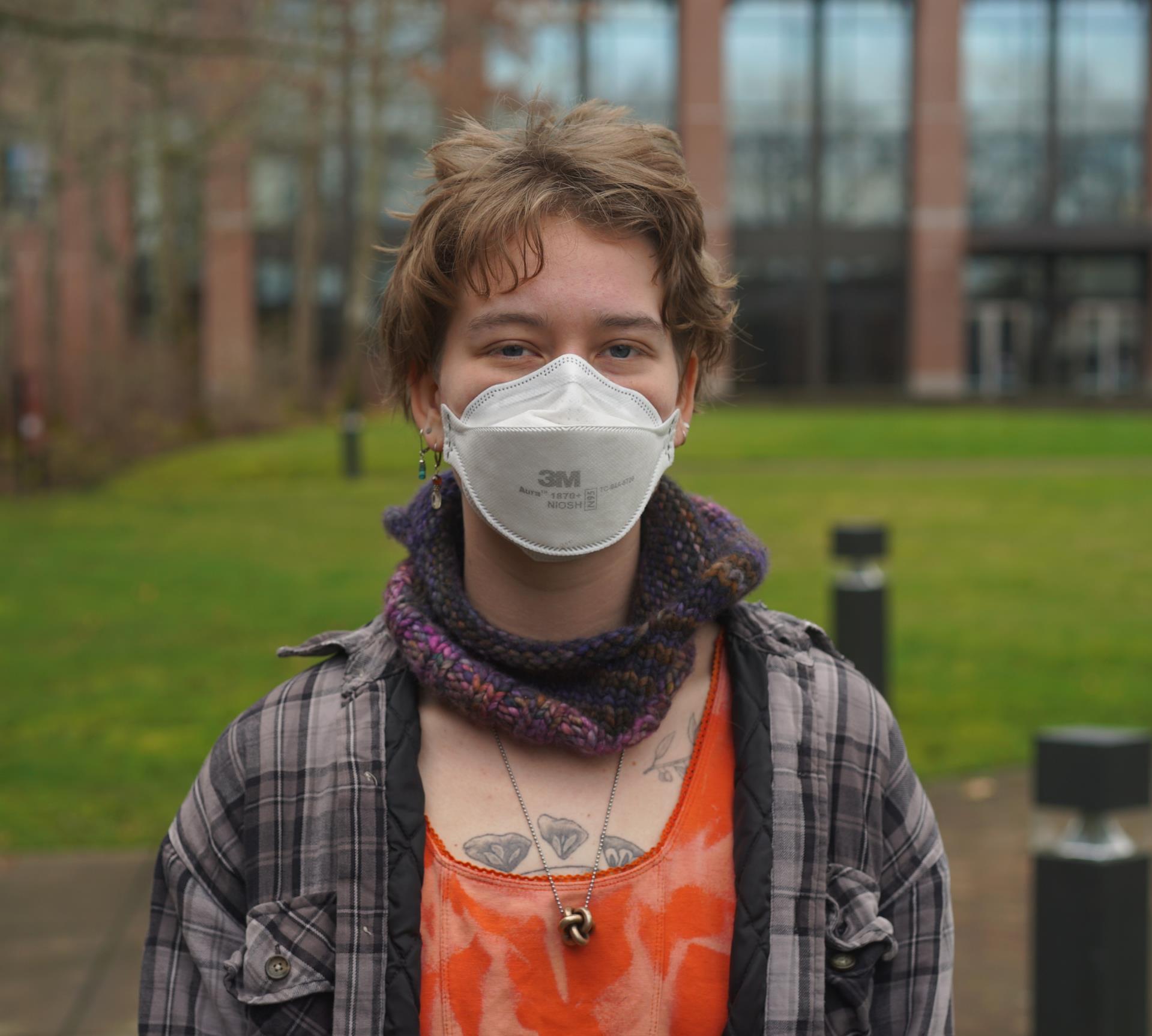 Morgan standing in front of Syre Student Center. They are wearing an orange top and flannel. They are wearing a surgical mask and have blonde hair