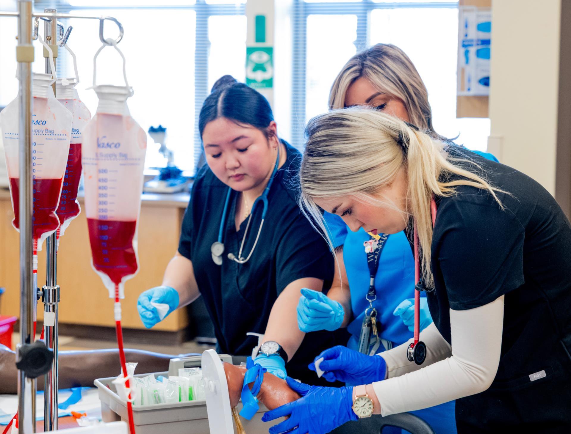Three Whatcom Community College Medical Assistant students in a lab drawing blood on a simulation lab hand.All students are wearing black scrubs.